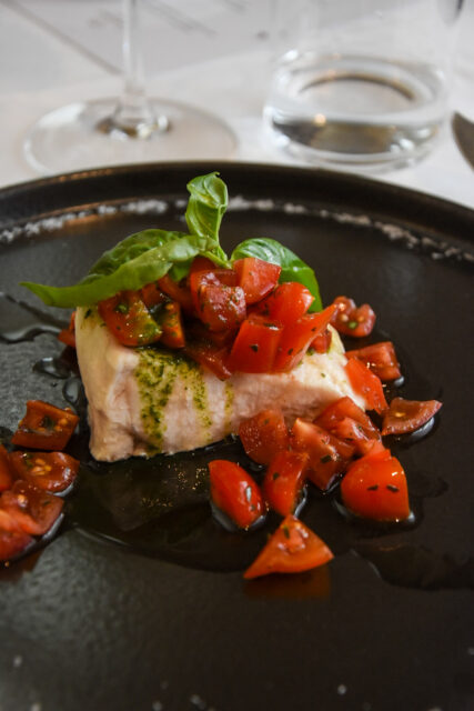 Un trancio di Carne di storione su un piatto ner oe coperto da pomodorini tagliati a pezzetti, pesto e qualche foglia di basilico