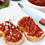 Due crostini con sopra la sardella spalmata. Su uno anche della cipolla rossa tritata.