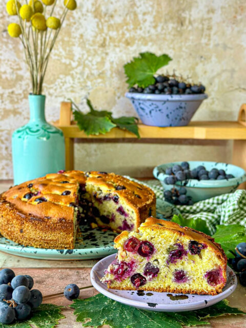 Una fetta di torta Bertolina con dietro la torta intera e un cestino di uva fragola