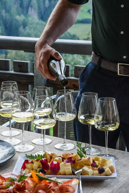 due piatti con salumi e formaggi e tanti calici di vino che una persona riempie con vino bianco